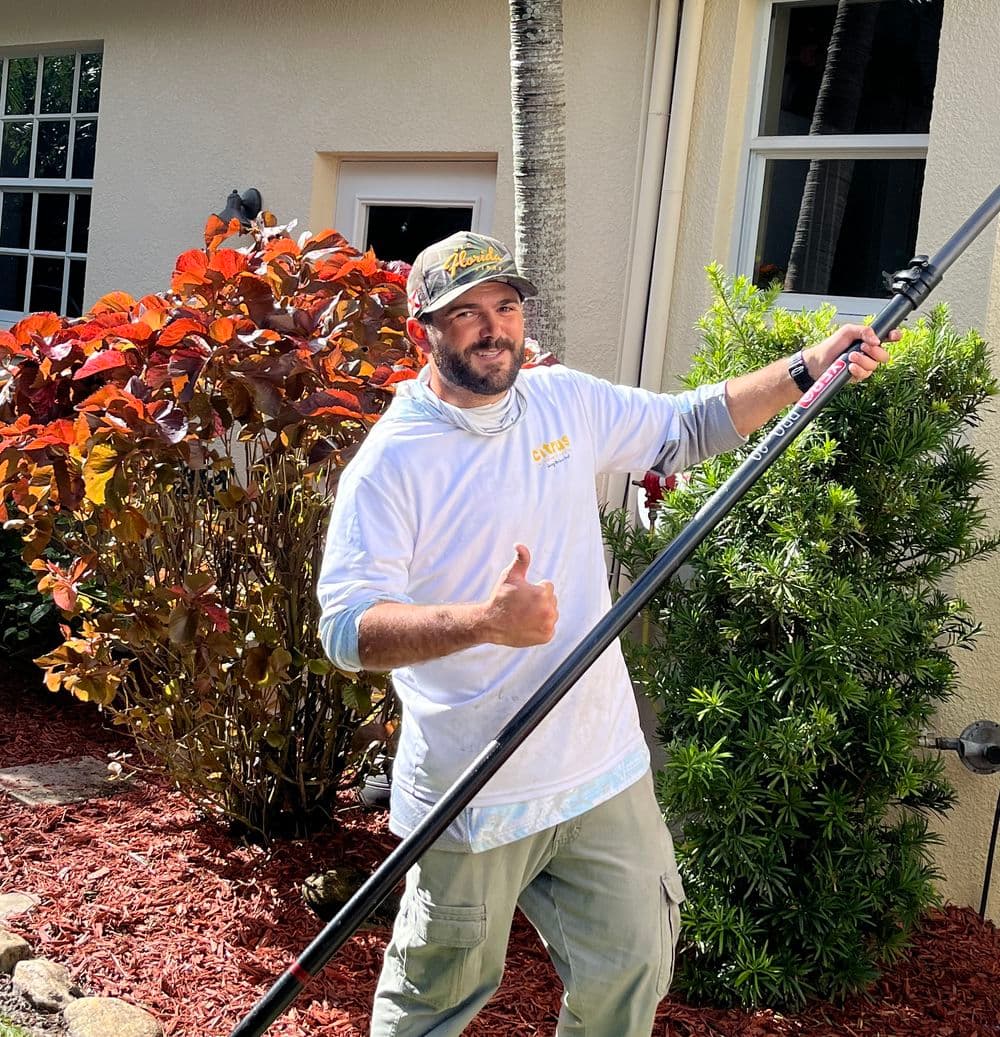 Man giving a thumbs up while holding a pole near colorful shrubs and a house exterior.