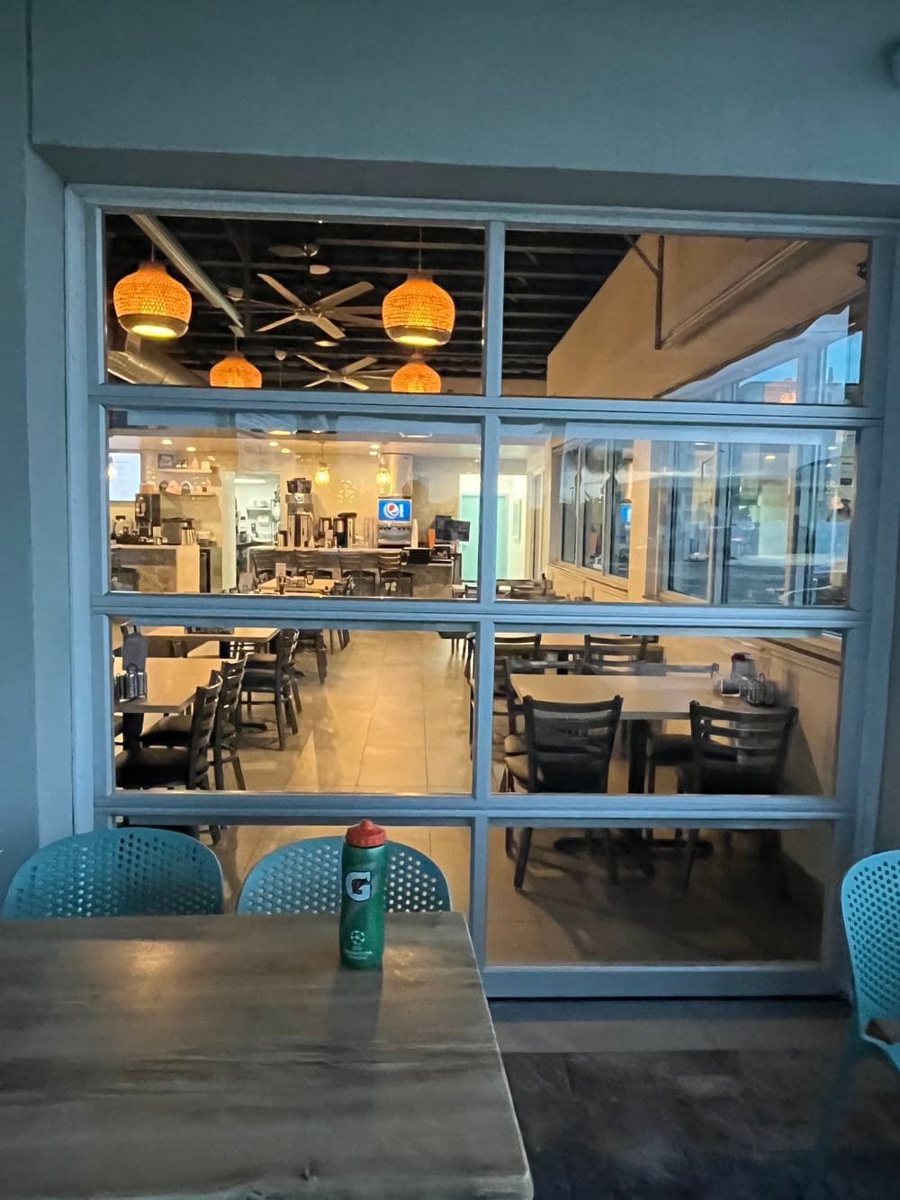 View of a modern restaurant interior with pendant lights and empty tables through glass windows.