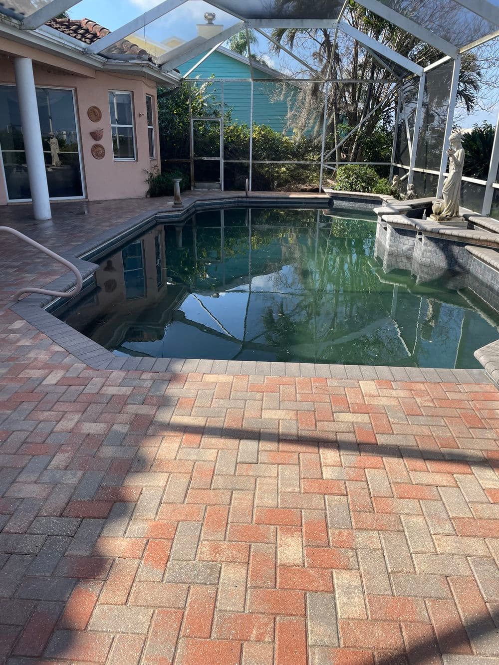 Elegant backyard pool area with paver deck, statue, and surrounding greenery under a screened enclosure.
