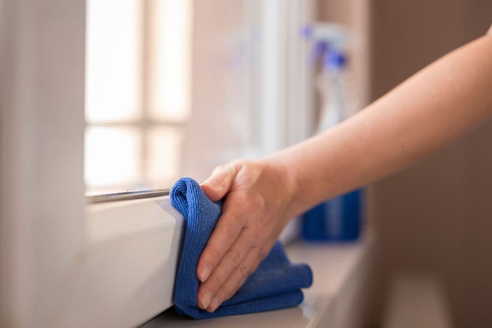 Person cleaning a window sill with a blue microfiber cloth and spray cleaner.